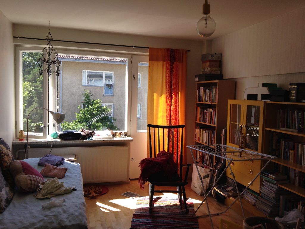 Kuvassa olohuone, takaseinällä ikkuna, josta tulee sisään valoa. Oikealla kirjahyllyt, vasemmalla vierasänky. Lattialla raidallinen matto, jonka päällä musta keinutuoli.